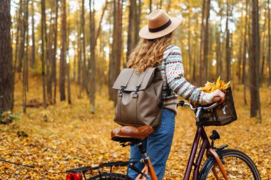 Parkta güneşli bir günde renkli sonbahar yapraklarıyla dolu bir bisikleti iterken gülümseyen genç bir kadın. Aktif turist güneşli havanın tadını çıkarıyor. Hafta sonu konsepti.