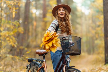 Parkta güneşli bir günde renkli sonbahar yapraklarıyla dolu bir bisikleti iterken gülümseyen genç bir kadın. Aktif turist güneşli havanın tadını çıkarıyor. Hafta sonu konsepti.