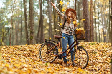 Parkta güneşli bir günde renkli sonbahar yapraklarıyla dolu bir bisikleti iterken gülümseyen genç bir kadın. Aktif turist güneşli havanın tadını çıkarıyor. Hafta sonu konsepti.