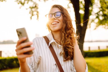 Kıvırcık saçlı güzel bir kadın, altın saat boyunca güneşli bir parkta akıllı telefonunu kullanırken gülümsüyor. Teknoloji, bağlantı, blog yazma,