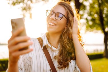 Kıvırcık saçlı güzel bir kadın, altın saat boyunca güneşli bir parkta akıllı telefonunu kullanırken gülümsüyor. Teknoloji, bağlantı, blog yazma,