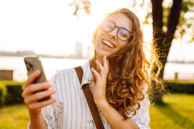 Kıvırcık saçlı güzel bir kadın, altın saat boyunca güneşli bir parkta akıllı telefonunu kullanırken gülümsüyor. Teknoloji, bağlantı, blog yazma,