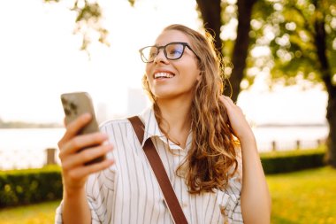 Kıvırcık saçlı güzel bir kadın, altın saat boyunca güneşli bir parkta akıllı telefonunu kullanırken gülümsüyor. Teknoloji, bağlantı, blog yazma,