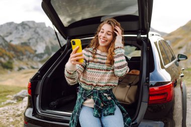 Genç bir gezgin kadın arabasının arkasında oturuyor, gülümsüyor ve telefonunu tepenin üstünde bulutlar asılı, manzaralı bir dağlık alanda kullanıyor. Seyahat, hafta sonu, doğa, rahatlama ve yaşam tarzı konsepti