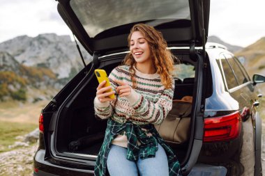 Genç bir gezgin kadın arabasının arkasında oturuyor, gülümsüyor ve telefonunu tepenin üstünde bulutlar asılı, manzaralı bir dağlık alanda kullanıyor. Seyahat, hafta sonu, doğa, rahatlama ve yaşam tarzı konsepti