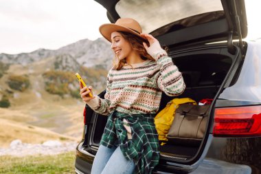 Genç bir gezgin kadın arabasının arkasında oturuyor, gülümsüyor ve telefonunu tepenin üstünde bulutlar asılı, manzaralı bir dağlık alanda kullanıyor. Seyahat, hafta sonu, doğa, rahatlama ve yaşam tarzı konsepti