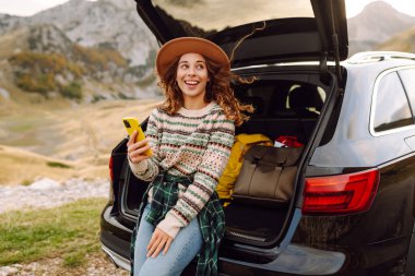 Genç bir gezgin kadın arabasının arkasında oturuyor, gülümsüyor ve telefonunu tepenin üstünde bulutlar asılı, manzaralı bir dağlık alanda kullanıyor. Seyahat, hafta sonu, doğa, rahatlama ve yaşam tarzı konsepti