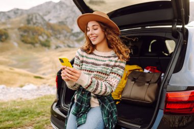 Genç bir gezgin kadın arabasının arkasında oturuyor, gülümsüyor ve telefonunu tepenin üstünde bulutlar asılı, manzaralı bir dağlık alanda kullanıyor. Seyahat, hafta sonu, doğa, rahatlama ve yaşam tarzı konsepti