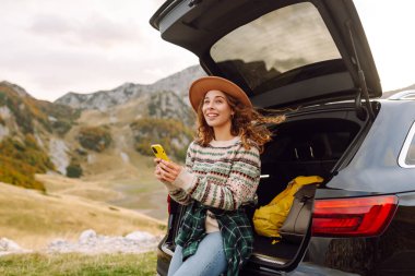 Genç bir gezgin kadın arabasının arkasında oturuyor, gülümsüyor ve telefonunu tepenin üstünde bulutlar asılı, manzaralı bir dağlık alanda kullanıyor. Seyahat, hafta sonu, doğa, rahatlama ve yaşam tarzı konsepti