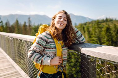 Genç bir gezgin kadın güneşli bir günde, dağlara tepeden bakan tahta bir köprüde durarak doğanın tadını çıkarıyor. Özgürlük kavramı, seyahat, yaşam tarzı. 