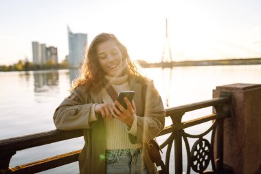 Nehir kenarında güneşli bir günün tadını çıkaran genç bir kadın, sıcak güneş ışığında gülümserken akıllı telefonunu kullanır.