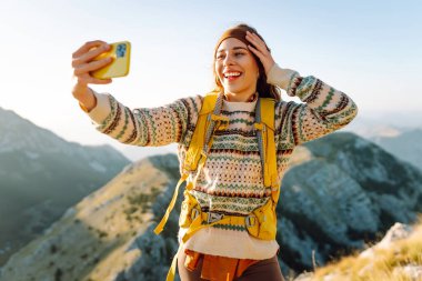 Güneşli bir günde dağ zirvesinde selfie çekerken yürüyüş macerasından zevk alan genç bir kadın. Doğa özgürlüğü macerası, mutlu tatiller. Günlük tutma.