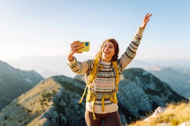 Güneşli bir günde dağ zirvesinde selfie çekerken yürüyüş macerasından zevk alan genç bir kadın. Doğa özgürlüğü macerası, mutlu tatiller. Günlük tutma.