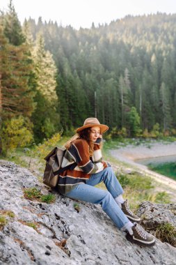 Güneşli bir günde ağaçlarla çevrili kayalık bir yüzeye oturmuş renkli bir kazak ve şapkayla doğanın tadını çıkaran genç bir gezgin kadın. Aktif hayat. Yaşam tarzı, seyahat, turizm