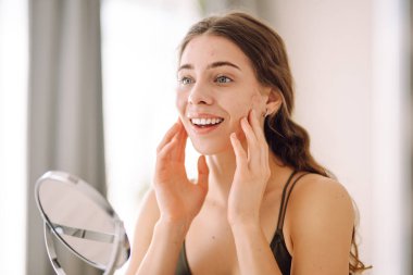 Derisi sorunlu genç bir kadın cilt bakımı rutiniyle meşguldür. İyi ışıklandırılmış, minimalist bir ortamda aynaya bakarken nazikçe yüzüne dokunur. Sivilce terapisi, yara, yüz iltihabı, sorunlu deri.. 