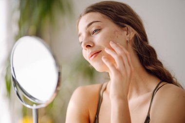 Derisi sorunlu genç bir kadın cilt bakımı rutiniyle meşguldür. İyi ışıklandırılmış, minimalist bir ortamda aynaya bakarken nazikçe yüzüne dokunur. Sivilce terapisi, yara, yüz iltihabı, sorunlu deri.. 