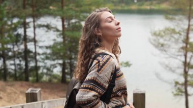 Genç bir kadın, göl kenarında huzurlu bir anın tadını çıkarırken kıtır bir sonbahar günü, doğal ortamda rahat bir kazak giyiyor.