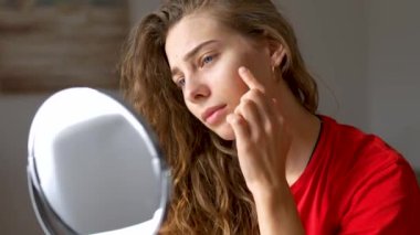 Güzellik ve doğallık kavramı. Genç bir kadın yüzündeki kızarıklıkları inceliyor. Aynaya bakarken tenine dokunuyor. Yüz bakımı. Sivilce.