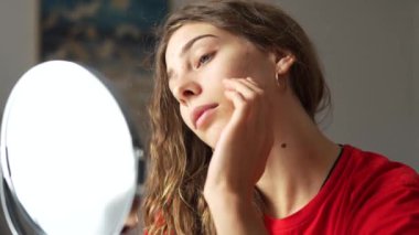 Güzellik ve doğallık kavramı. Genç bir kadın yüzündeki kızarıklıkları inceliyor. Aynaya bakarken tenine dokunuyor. Yüz bakımı. Sivilce.