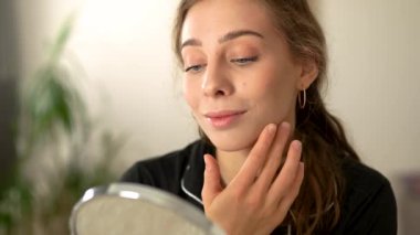 Genç bir kadın aynadaki yansımasına hayran, yüz derisini inceliyor. Mutlu kadın, aydınlık odada yüz bakımı için eğleniyor. Güzellik, doğallık konsepti.