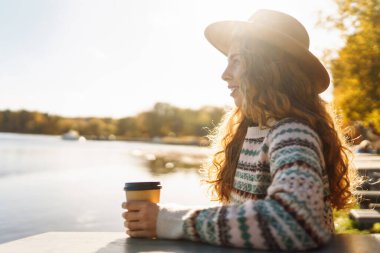 Nehrin kenarında oturan, elinde kahvesiyle genç bir kadın. Sonbahar Sabahı.