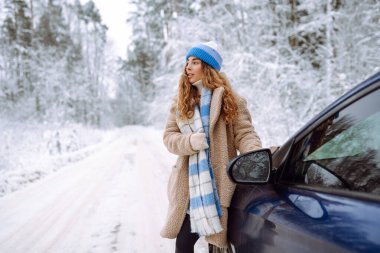 Kışın arabasının yanında güzel genç bir kadın ilk karın tadını çıkarıyor. Kış Sessizliği. Bir gezgin doğada bir arabanın yanında poz verir. Araba yolculuğu, tatil kavramı.