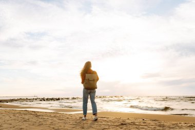 O, plaj boyunca yürürken, kadın turist sırt çantasını ayarladı, tuzlu deniz havasını ve gün batımında dalgaların sesini içine aldı. Sakinlik ve sükunet. Seyahat, turizm konsepti.