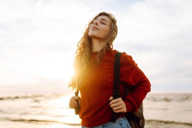 O, plaj boyunca yürürken, kadın turist sırt çantasını ayarladı, tuzlu deniz havasını ve gün batımında dalgaların sesini içine aldı. Sakinlik ve sükunet. Seyahat, turizm konsepti.