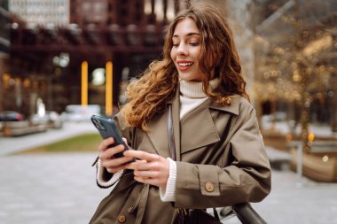 Trençkotlu çekici bir kadın sokakta telefon kullanıyor. Yaşam tarzı, seyahat, turizm, teknoloji