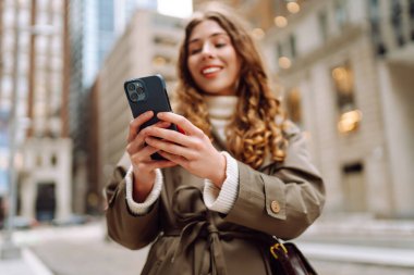 Trençkotlu çekici bir kadın sokakta telefon kullanıyor. Yaşam tarzı, seyahat, turizm, teknoloji