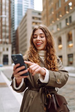 Trençkotlu çekici bir kadın sokakta telefon kullanıyor. Yaşam tarzı, seyahat, turizm, teknoloji