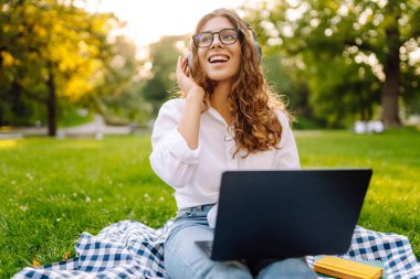 Yaz günü dizüstü bilgisayar ve kablosuz kulaklık kullanırken bacak bacak üstüne atmış yeşil çimlerde oturan çekici genç bir kadının portresi. Eğitim, iş, blog veya serbest çalışma kavramı.