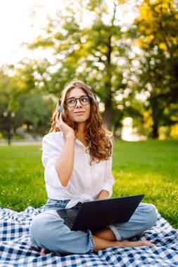 Yaz günü dizüstü bilgisayar ve kablosuz kulaklık kullanırken bacak bacak üstüne atmış yeşil çimlerde oturan çekici genç bir kadının portresi. Eğitim, iş, blog veya serbest çalışma kavramı.