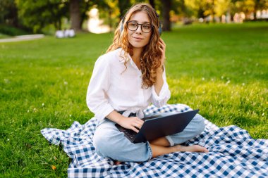 Yaz günü dizüstü bilgisayar ve kablosuz kulaklık kullanırken bacak bacak üstüne atmış yeşil çimlerde oturan çekici genç bir kadının portresi. Eğitim, iş, blog veya serbest çalışma kavramı.