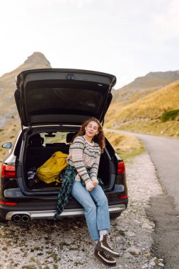 Genç bir kadın arabasının bagajında oturuyor, kayalık araziden geçen virajlı bir yolun kenarına park etmiş. Yolculuklar. Sonbahar manzarası