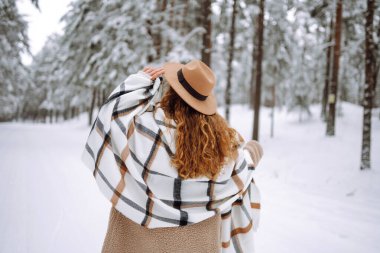 Kış ormanlarında karlı ağaçların arasında dikilen ve ilk karın tadını çıkaran kadının arka görüntüsü. Şapka, ekose atkı ve palto giyiyor. Gezgin şehveti ve boho stili