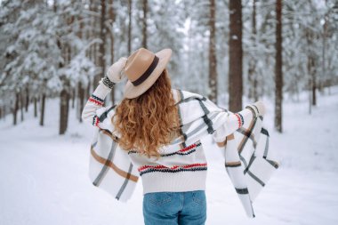 Kış ormanlarında karlı ağaçların arasında dikilen ve ilk karın tadını çıkaran kadının arka görüntüsü. Şapka, ekose atkı ve palto giyiyor. Gezgin şehveti ve boho stili