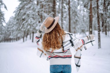 Kış ormanlarında karlı ağaçların arasında dikilen ve ilk karın tadını çıkaran kadının arka görüntüsü. Şapka, ekose atkı ve palto giyiyor. Gezgin şehveti ve boho stili