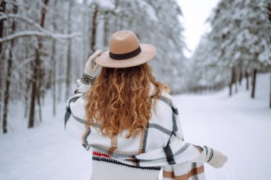 Kış ormanlarında karlı ağaçların arasında dikilen ve ilk karın tadını çıkaran kadının arka görüntüsü. Şapka, ekose atkı ve palto giyiyor. Gezgin şehveti ve boho stili