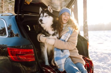 Young woman in winter clothes sitting in the trunk of a car with her husky and play. Winter holidays. Travel concept