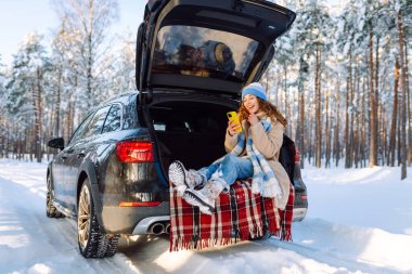 Bagajında oturan ve kış vakti telefon kullanan genç bir kadın. Teknoloji kavramı, macera, arabayla seyahat..