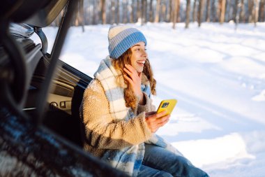 Bagajında oturan ve kış vakti telefon kullanan genç bir kadın. Teknoloji kavramı, macera, arabayla seyahat..