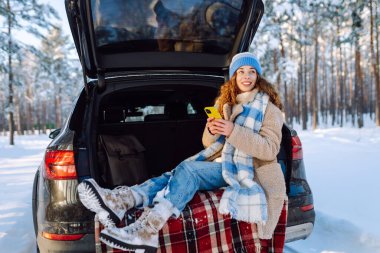 Bagajında oturan ve kış vakti telefon kullanan genç bir kadın. Teknoloji kavramı, macera, arabayla seyahat..