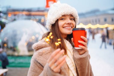 Gülümseyen kadın elinde sıcak kahveyle bir Noel pazarında yürüyor. Noel festivali, kış tatili konsepti..