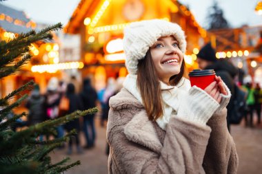 Gülümseyen kadın elinde sıcak kahveyle bir Noel pazarında yürüyor. Noel festivali, kış tatili konsepti..