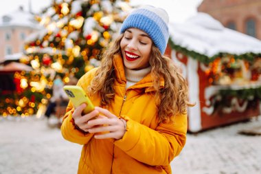 Arka planda dekoratif Noel ışıkları olan akıllı telefonu kullanan genç bir kadın. Noel Fuarı.