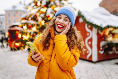 Arka planda dekoratif Noel ışıkları olan akıllı telefonu kullanan genç bir kadın. Noel Fuarı.