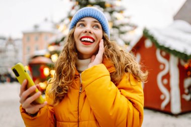 Arka planda dekoratif Noel ışıkları olan akıllı telefonu kullanan genç bir kadın. Noel Fuarı.