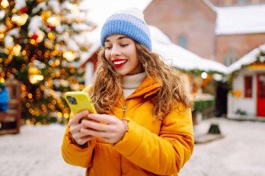 Arka planda dekoratif Noel ışıkları olan akıllı telefonu kullanan genç bir kadın. Noel Fuarı.