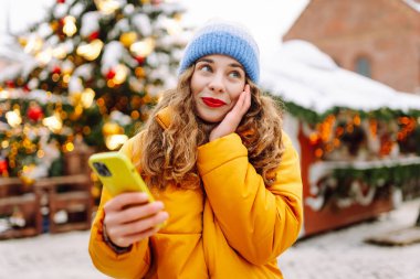 Arka planda dekoratif Noel ışıkları olan akıllı telefonu kullanan genç bir kadın. Noel Fuarı.